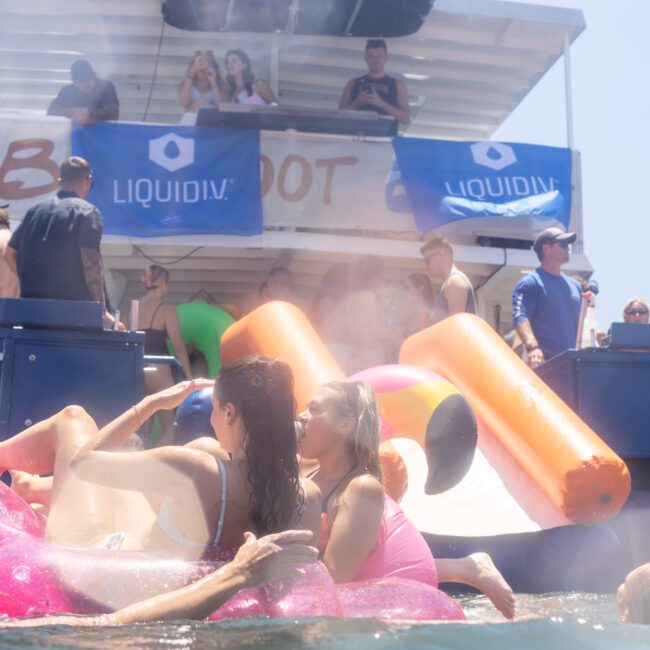 A group of people enjoying a party on a boat with inflatable floats in the water. Some are sitting on large inflatable rings, while others are on the boat, under a blue sky. Banners with logos are visible on the boat.