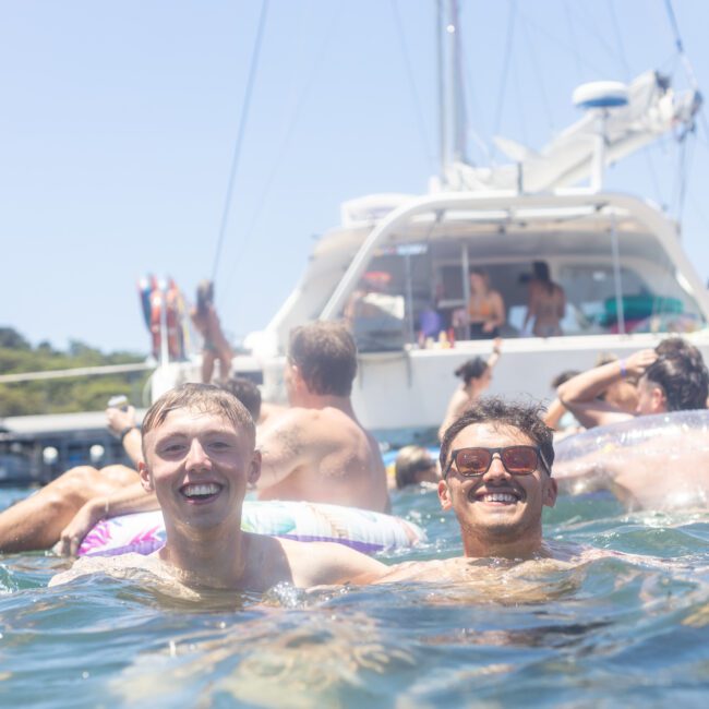 Two people smiling in the water near a boat. The boat is crowded with people relaxing and sunbathing. The sky is clear, and there are inflatable floats around. The scene suggests a lively day of fun on the water.