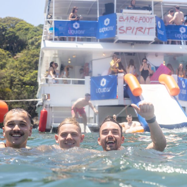 Three people are smiling in the water near a large boat decorated with banners reading "Barefoot Spirit." The boat has people on board and a slide extending into the water. The setting is a sunny, outdoor scene.