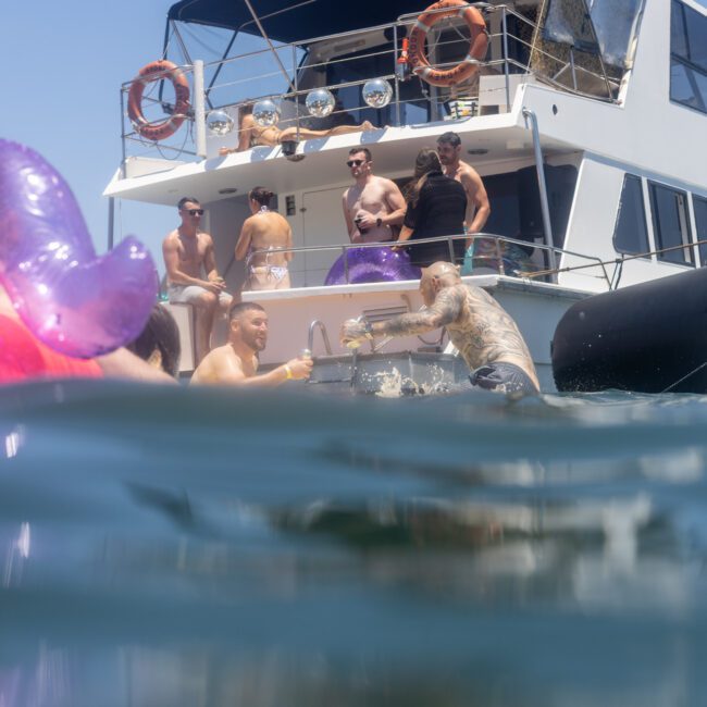 A group of people enjoying a sunny day on a yacht. Some are in the water with inflatable floats, while others are on the deck. The sky is clear and the atmosphere is lively and relaxed.