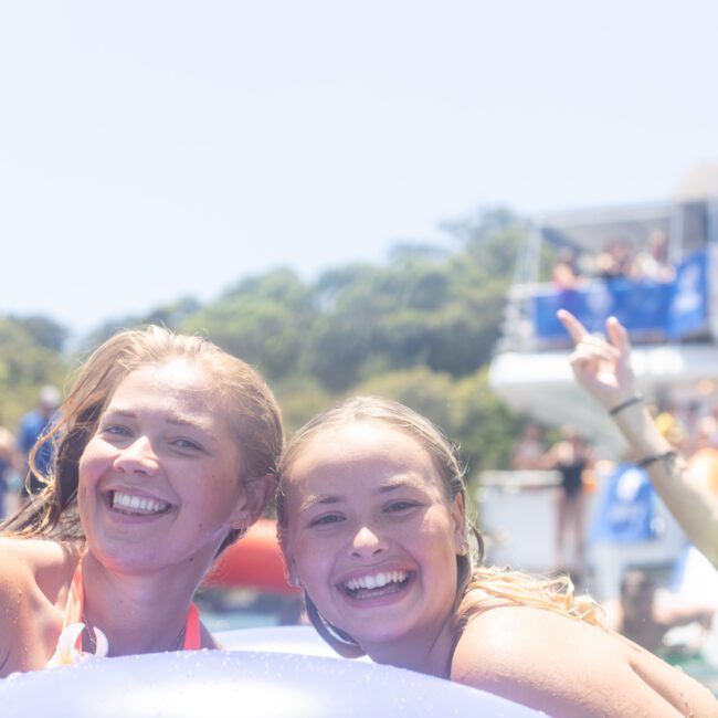 Two smiling women in swimsuits enjoy a sunny day at a pool event. People in the background are on a boat, with one person raising a peace sign. The atmosphere is festive and bright.