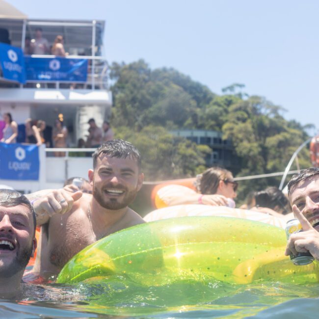 Three men are smiling and enjoying themselves in the water, holding a green inflatable float. A boat with people on it is in the background, and trees can be seen on the shore. The atmosphere is lively and festive.