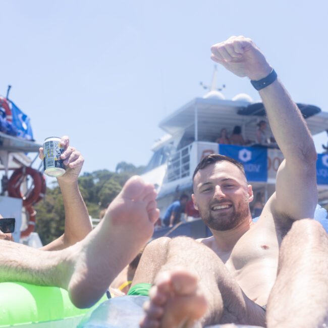 Two men relax on inflatable floats, smiling and enjoying drinks on a sunny day. A boat with people in the background suggests a lively gathering. One man raises his arm in celebration or greeting.