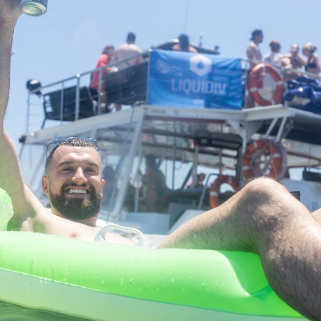 A man smiles and relaxes on a green float in the water, holding a drink can. In the background, a group of people are gathered on a boat with a blue "LIQUIDIV" banner. The sky is clear and sunny.