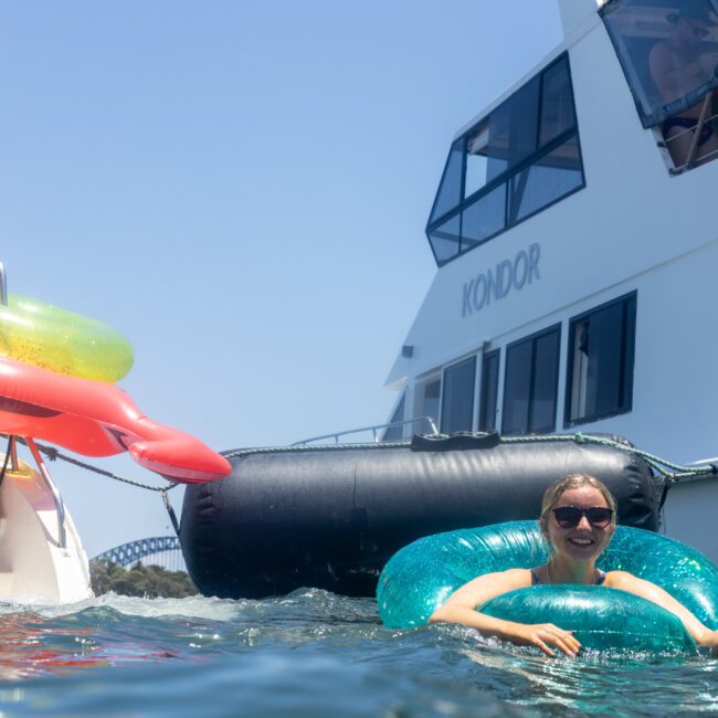 A person is enjoying the water while floating on an inflatable tube near a yacht. The yacht is docked and has multiple inflatables on board. The sky is clear and sunny.