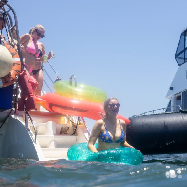 Two people enjoying a sunny day on a yacht. One person is standing by the yacht's steps with drinks and the other is in the water with a green inflatable ring. Bright inflatable floats are visible in the background.