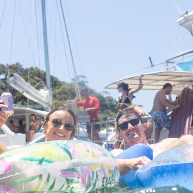 A man and a woman relax on inflatable pool floats in the water, smiling and enjoying a sunny day. The woman holds a can, and boats with other people are visible in the background.