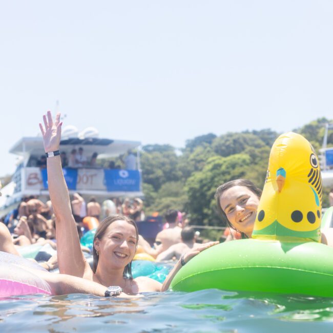 People enjoying a sunny day in a pool with flotation devices, including an inflatable duck. A boat and trees are visible in the background. One person waves at the camera, smiling.