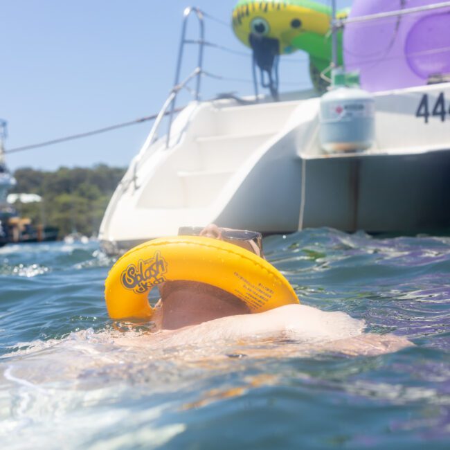 A person wearing a yellow floating device and goggles is swimming near a boat. The water is clear and the sky is blue. The boat's registration number is visible, and there are inflatables on board. Other boats are in the background.