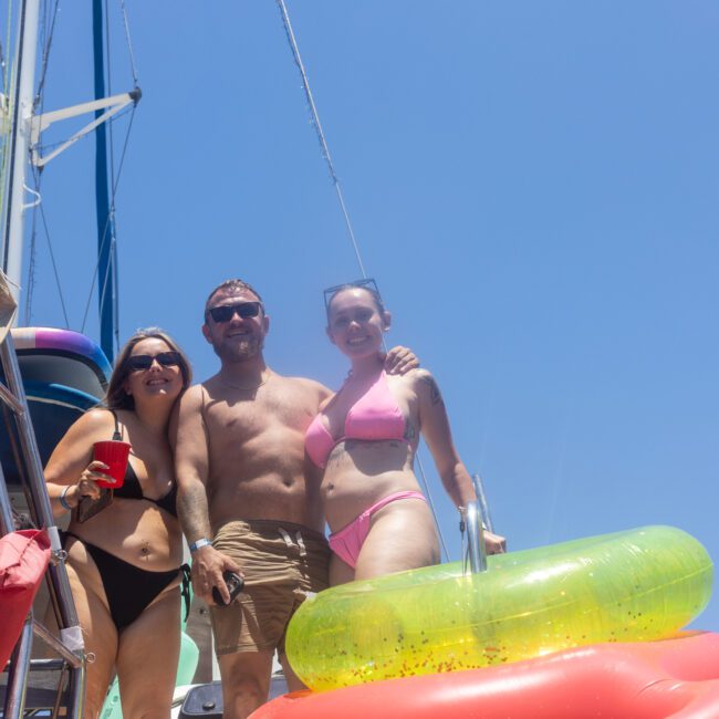 Three people in swimwear pose on a boat under a clear blue sky. They are surrounded by colorful inflatable pool floats. One person holds a red cup.