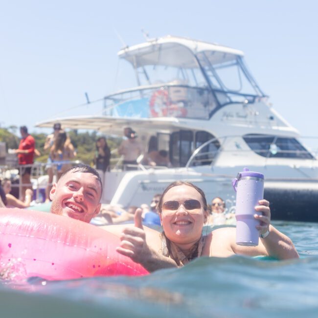 Two people smiling and giving thumbs up while floating on an inflatable raft in the water. A boat with people on it is in the background, and the scene is sunny and lively.