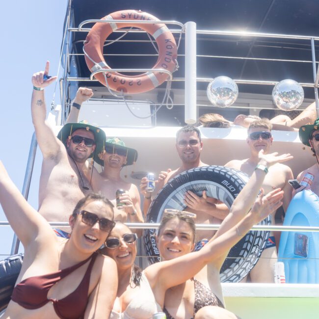 A group of people on a boat, smiling and posing with inflatable rings. They are wearing swimsuits and sunglasses. The sun is shining, and there are life rings and silver disco balls in the background.