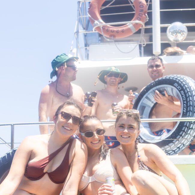 A group of people enjoying a sunny day on a boat. Three women in the foreground are smiling at the camera, wearing swimsuits and sunglasses. Several men are in the background, some wearing hats and holding inflatable toys.