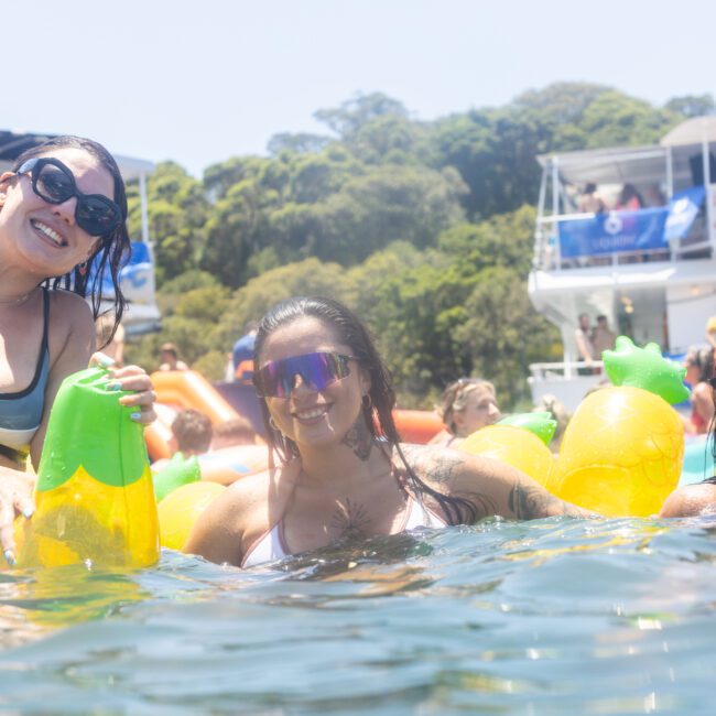 Three people in swimsuits and sunglasses enjoy the water, holding inflatable toys shaped like pineapples. Boats with passengers are in the background, suggesting a lively, sunny day on the water.