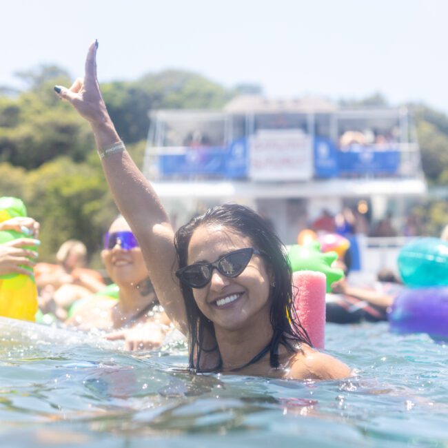 A woman in sunglasses smiles while standing in a pool, raising one hand in the air. She is surrounded by other people enjoying the water, and a boat is visible in the background.