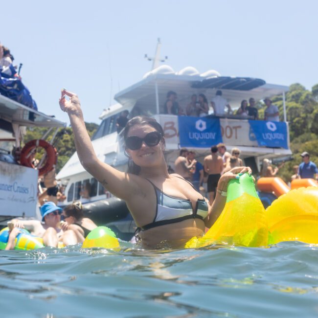 A woman in a bikini, smiling and raising her arm, floats on a yellow inflatable in the water. Behind her, a crowded boat with people enjoying a sunny day. The background features lush greenery and a clear blue sky.