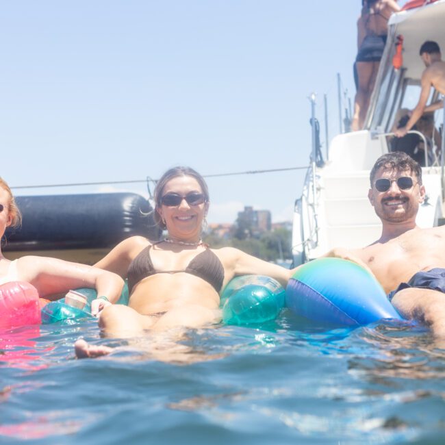 Three people lounging on colorful inflatables in the water, smiling at the camera. A yacht is in the background with more people enjoying the sunny day.