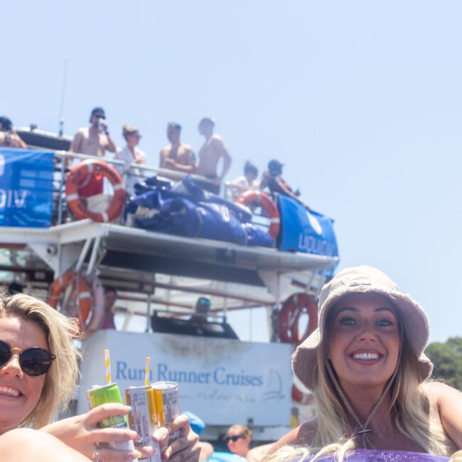 Two women smile while holding drinks on inflatable rafts. Behind them is a large boat with people on the upper deck. The boat has banners with logos and text that reads "Run Runner Cruises." The sky is clear and sunny.