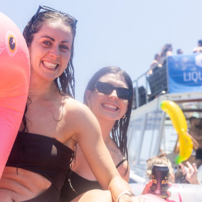 Two women in swimsuits smile while sitting on a pink inflatable flamingo. They're surrounded by others enjoying a sunny day on a boat.