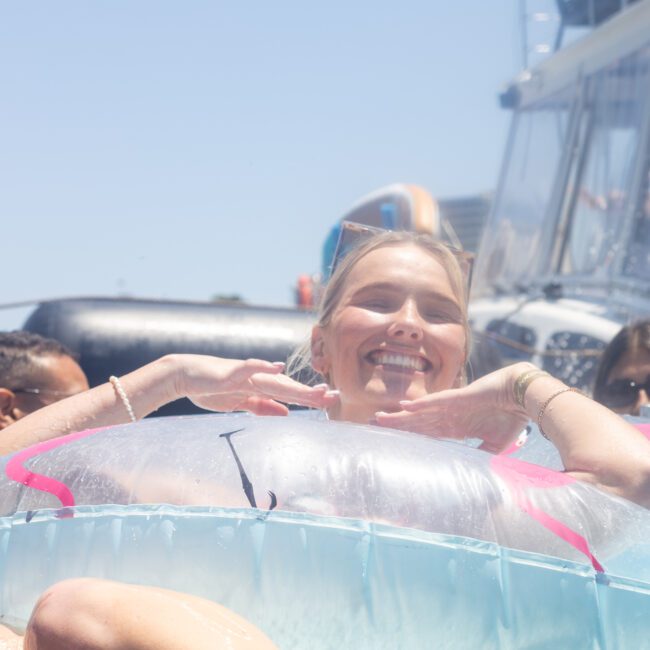 A person smiling and relaxing on a large inflatable float in a body of water, surrounded by other people and boats under a sunny sky.