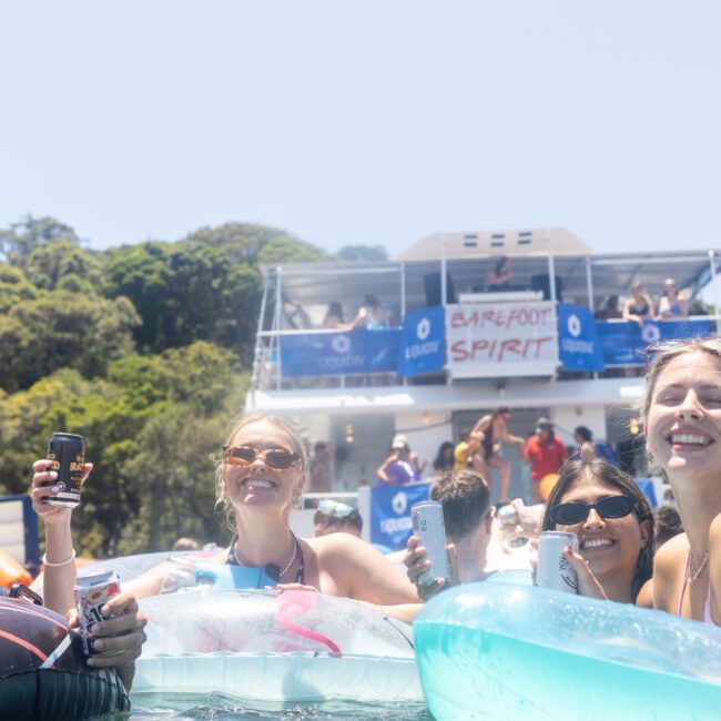 A group of people are smiling and relaxing on inflatable rafts in the water near a boat named "Barefoot Spirit." Some are holding drinks. The boat is anchored near a forested shoreline under a clear sky.