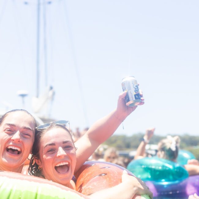 Two people smiling and laughing in a pool, each holding up a drink can. They are surrounded by colorful inflatable floaties. The background shows blurred boats and more people enjoying the sunny day.