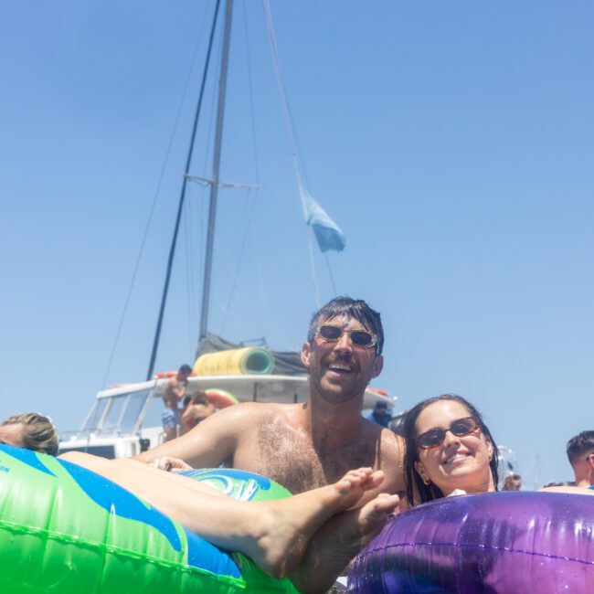 A group of people are enjoying themselves in a pool while floating on colorful inflatable tubes. A boat with sails is visible in the background under a bright, clear blue sky. Everyone appears relaxed and happy.