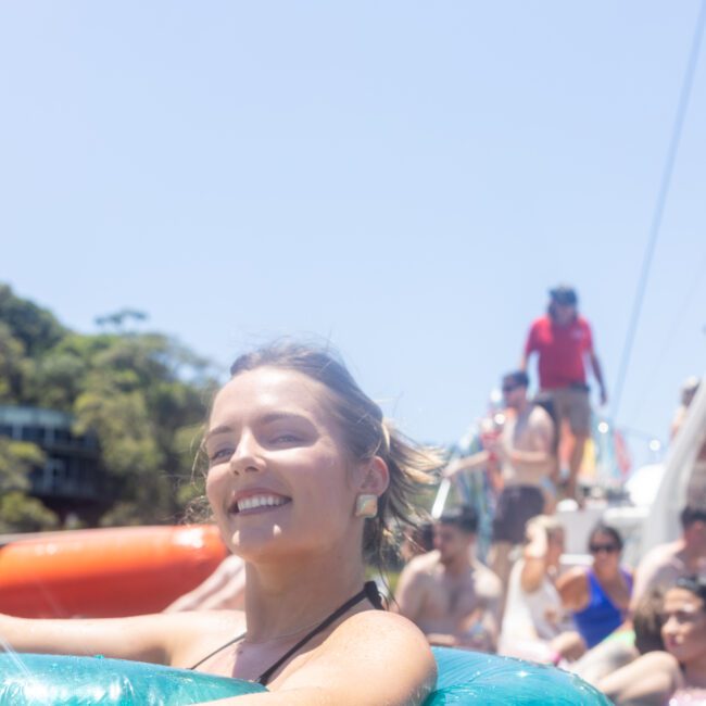 A smiling person in a blue inflatable ring enjoys a sunny day on the water, surrounded by a group of people on a boat in the background. The sky is clear, and it appears to be a lively and relaxing atmosphere.