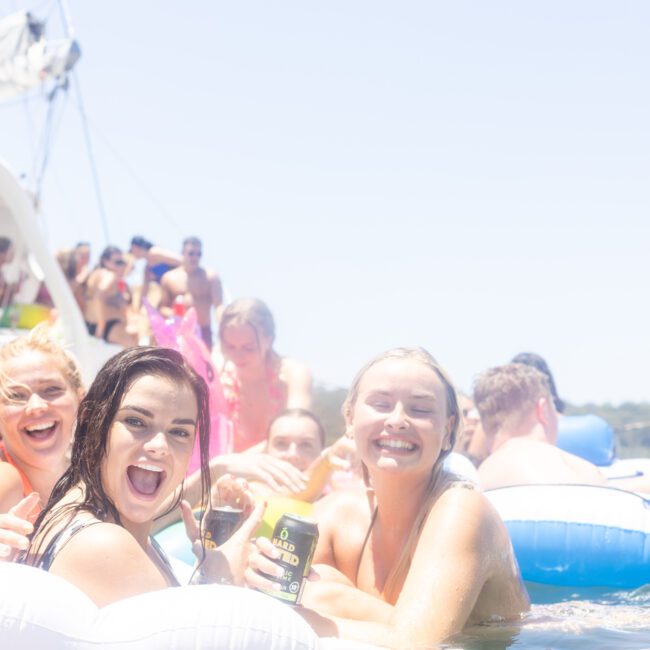 A group of people having fun in the water near a boat. They are smiling, holding drinks, and sitting on inflatable floats. The scene is bright and sunny, suggesting a festive atmosphere.