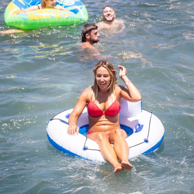 A woman in a red bikini floats on a blue and white inflatable ring in the water. Two men swim nearby, and another person is on a green inflatable in the background. It's a sunny day.