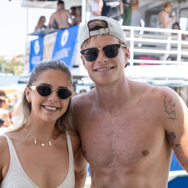 A smiling woman and man wearing sunglasses pose together on a boat. The man is shirtless with a tattoo on his arm, and the woman is in a light swimsuit. Other people and boats are in the background.