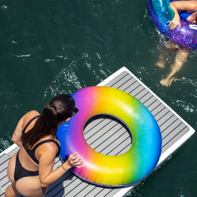 A woman in a black swimsuit stands on a dock holding a rainbow-colored inflatable ring. Another woman floats in a purple galaxy-patterned ring in the water. The scene is sunny and vibrant.