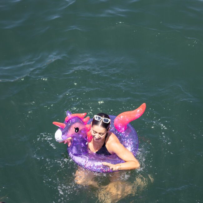 A person in sunglasses enjoys the water, floating with a purple, unicorn-shaped inflatable ring. The clear green-blue water surrounds them as they smile, with a watermark in the bottom right corner.