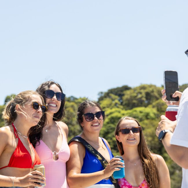 A group of people pose for a photo outdoors under a clear blue sky. Four individuals stand together smiling, wearing swimsuits and sunglasses, holding drinks. Another person in a white shirt holds a phone, capturing the moment. Trees are in the background.