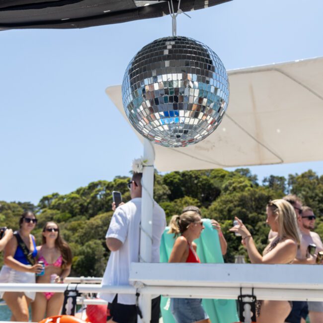 A group of people enjoying a boat party on a sunny day. There's a shiny disco ball hanging above them. Some people are taking photos, while others are chatting. Lush greenery is visible in the background.