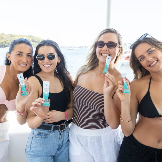 Four women smiling and holding packets while standing on a boat. They are wearing swimwear and sunglasses. The background shows water and trees in the distance, suggesting a sunny day.