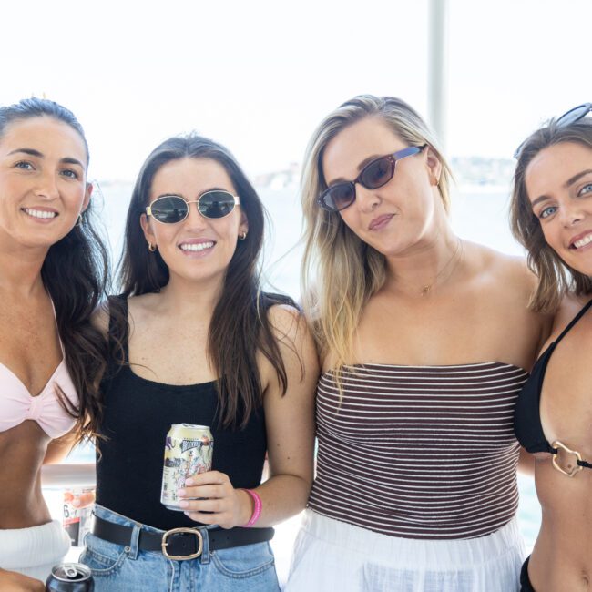 Four women in swimsuits and sunglasses smiling at the camera. One is holding a drink can. They appear to be outdoors near water, enjoying a sunny day.