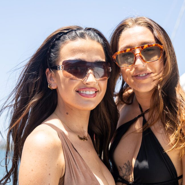 Two women in sunglasses smile at the camera on a sunny day. They wear swimsuits and stand on a boat with water and a clear blue sky in the background.