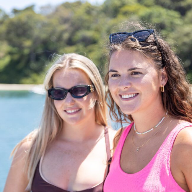 Two women are smiling outdoors with a scenic background of water and trees. One wears a brown top and sunglasses, the other a pink top and sunglasses on her head. It's a sunny day.