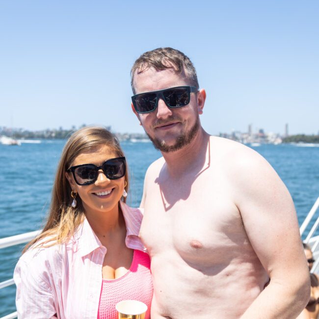 A man and woman, both wearing sunglasses, pose on a boat under a clear blue sky. The woman wears a pink shirt and holds a cup, while the shirtless man stands beside her. The ocean and cityscape are visible in the background.