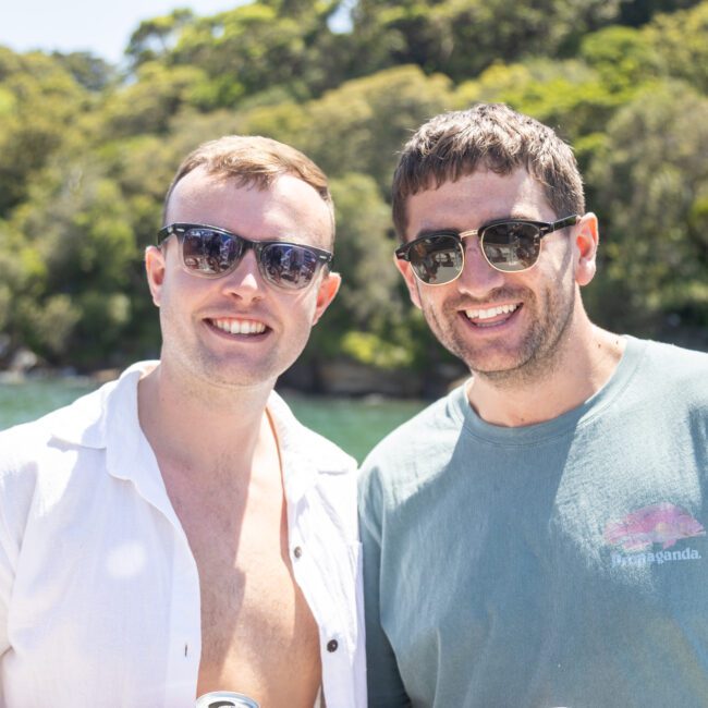 Two men wearing sunglasses smile while standing outdoors. One wears a white shirt and the other a green t-shirt. They are in a sunny setting with greenery and water in the background.