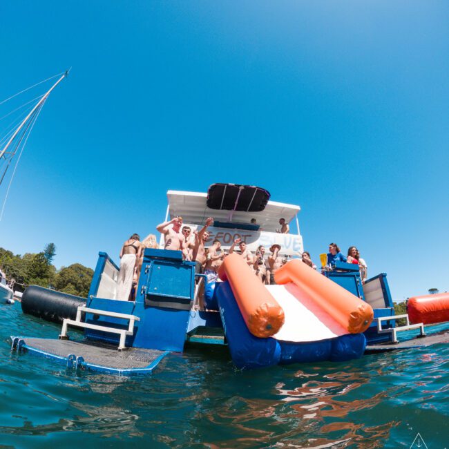 A group of people enjoying a sunny day on a floating platform in the water, featuring inflatable slides and lounges. The platform is colorful with blue, red, and orange elements. Clear sky and distant trees are visible.