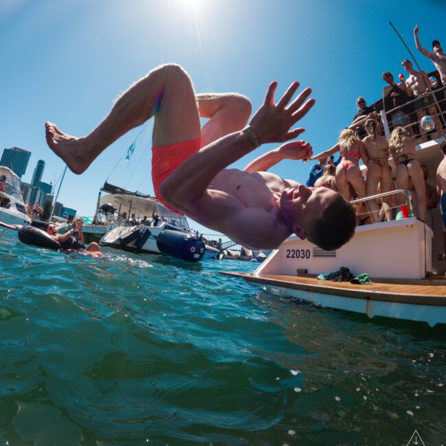 A person in red swim trunks is captured mid-air doing a backflip off a boat into the water. Several people watch from the deck under a clear blue sky, with city buildings visible in the background.