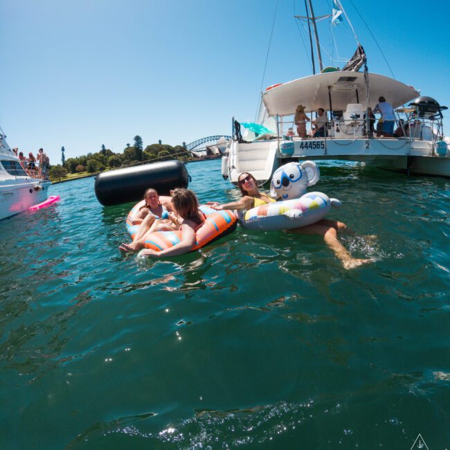 People relaxing on colorful inflatable tubes in the water near a docked sailboat. The background features more boats and an arched bridge under a clear blue sky.