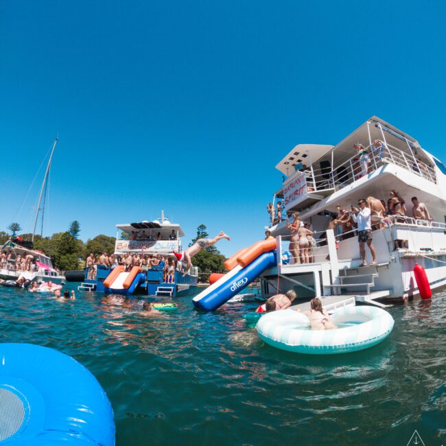 A group of people enjoying a sunny day on a lake with inflatable slides and pool floats. Two large boats are anchored close by, and some people are on the water. The sky is clear and blue, creating a vibrant summertime scene.