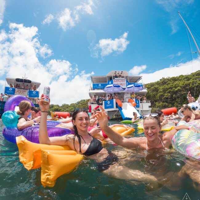 Two people in swimsuits enjoying a party on inflatable floats in the water. They are holding hands and drinks, surrounded by other partygoers and boats. It’s a sunny day with blue skies and fluffy clouds.