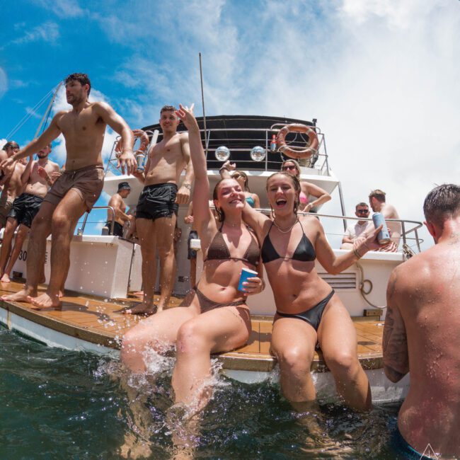 A group of people in swimsuits enjoy a sunny day on a boat. Two women sit on the boat's edge with feet in the water, smiling and holding cups. Others stand or sit in the background, with blue skies overhead.