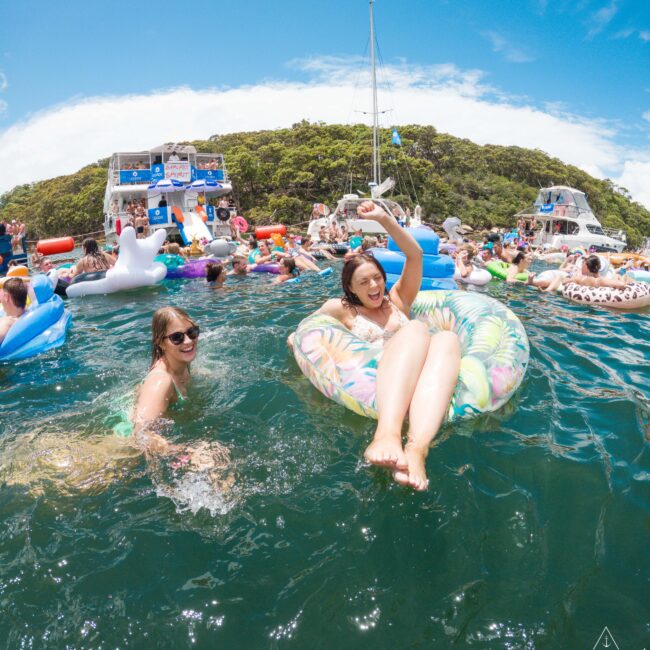 A woman in a floral inflatable ring and another in a pool float in blue-green water. They're surrounded by a crowd enjoying a sunny day with boats and lush hills in the background. The atmosphere is lively and festive.