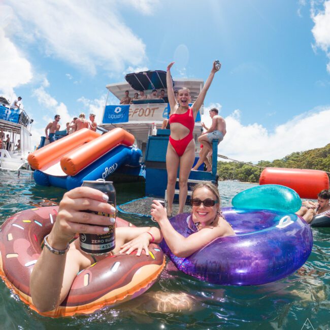 A joyful group of people having fun at a floating pool party. One person stands on a platform in a red swimsuit, raising hands. Others relax in colorful inflatable rings, one holding a drink. A boat and lush greenery are in the background under a clear sky.