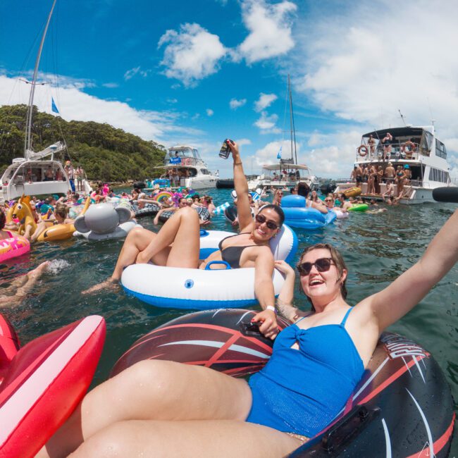 People floating on inflatable tubes in a lively gathering on the water, with boats and a crowd in the background. Two individuals in the foreground are smiling and holding drinks, enjoying a sunny day at a water party.